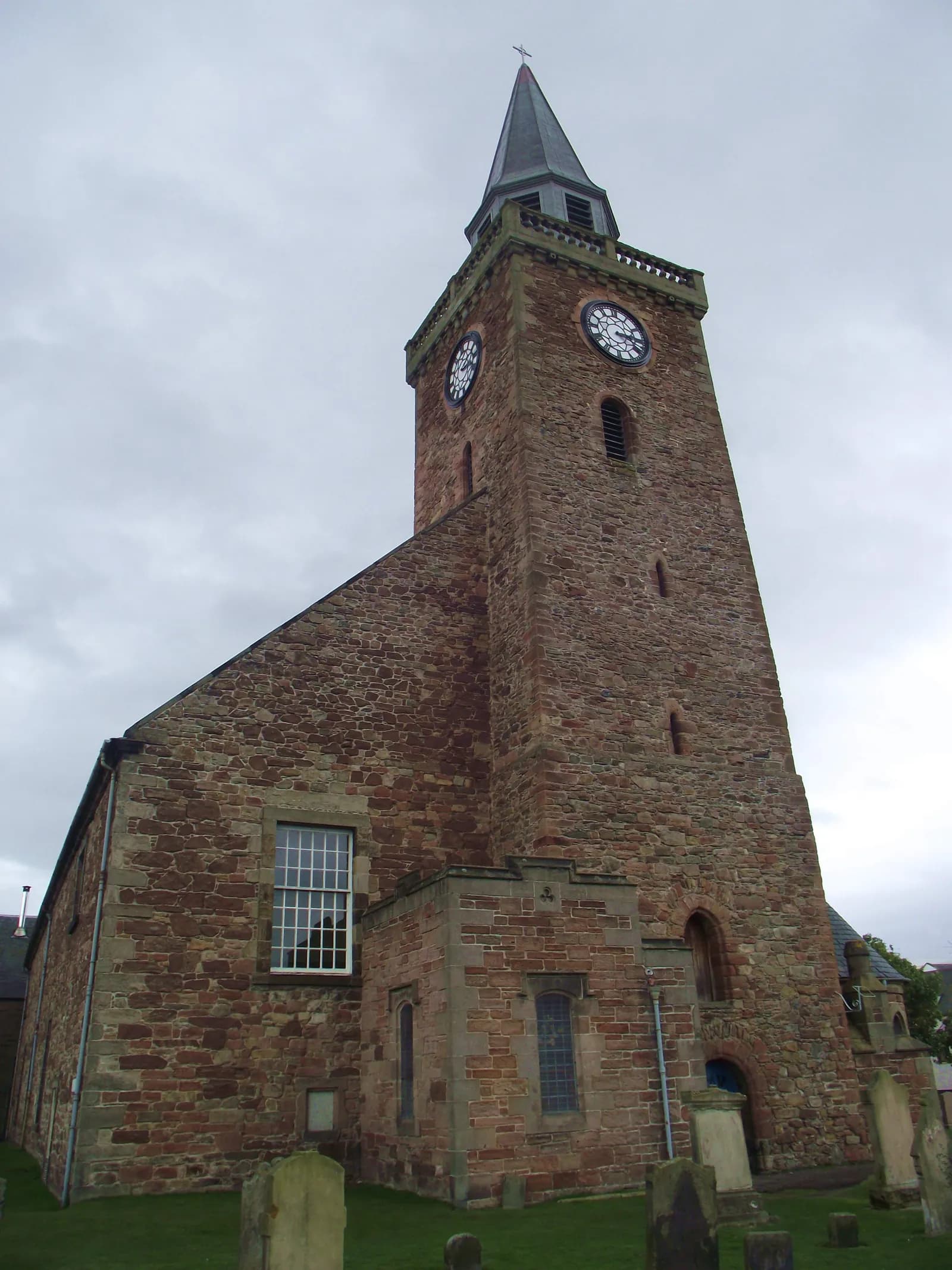 A historic stone church with a tall clock tower, set against a cloudy sky, with gravestones visible in the grassy foreground.